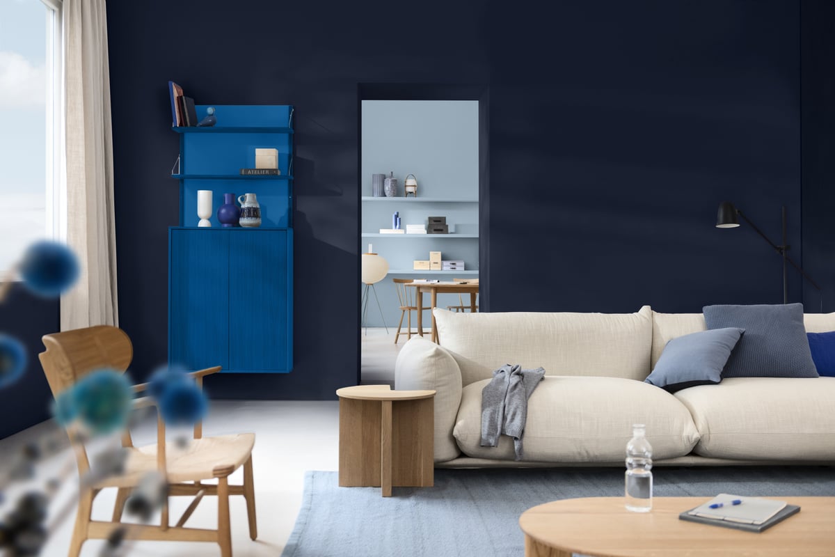 Woonkamer met donkerblauwe muur, crèmekleurige bank en blauwe kast; in de deuropening staat een eettafel.