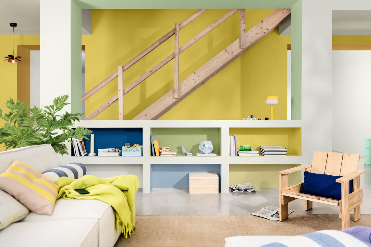 Woonkamer met gele trapwand, open vakkenkast, witte bank met groene plaid en houten stoel met blauw kussen.