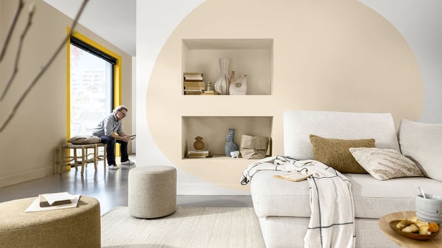 Helder woonkamer met beige muur, planken en bank; op de achtergrond werkt een persoon bij het raam.