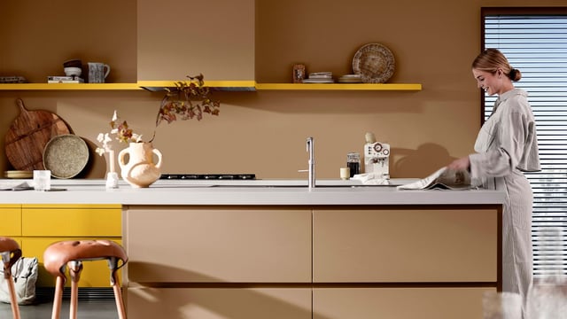 Vrouw veegt het aanrecht in een moderne keuken met gele accenten en planten op het plankje.
