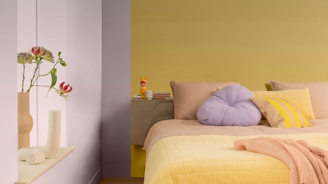 Lichte slaapkamer met bed, kussens en een vaas met bloemen op een plank.