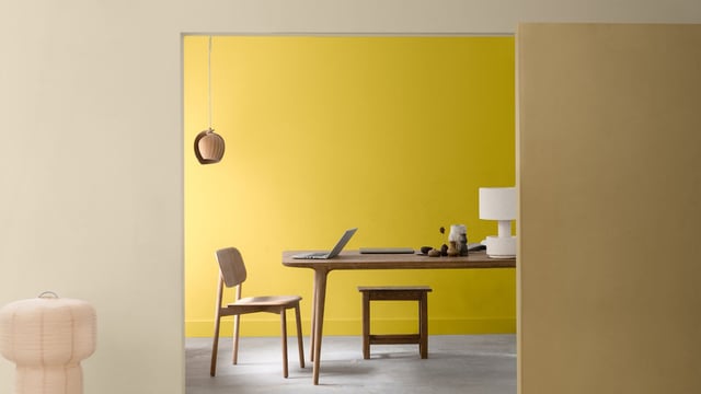 Licht thuiswerkplek met gele accentmuur, bureau, stoelen en hanglamp.