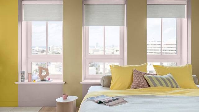 Lichte slaapkamer met gele accentmuur, bed met gele beddengoed en grote ramen met jaloezieën.