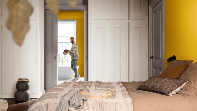 Lichte slaapkamer met bed en ingebouwde kasten; een man loopt door de deuropening met een knuffel.