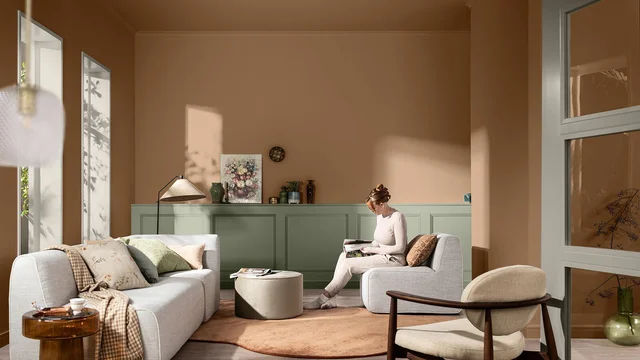 Gezellige woonkamer met bank, fauteuil, salontafel, open boeken en een vrouw die leest bij daglicht.