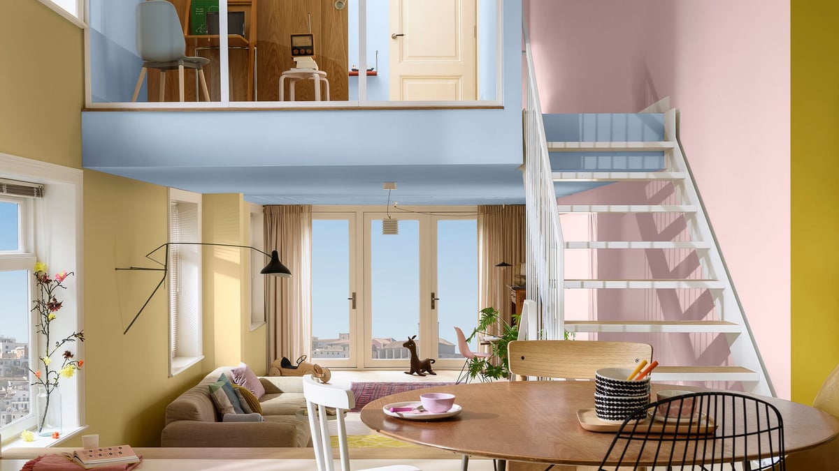 Zonnige loftwoonkamer met pastelwanden, witte trap, eettafel op de voorgrond en bank bij grote ramen.